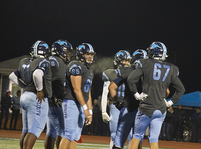 A group of offensive linemen gathering together during a timeout. The temperature was cold enough to see the breath coming from their mouth and the steam coming off of the players jerseys while they were sweating. 